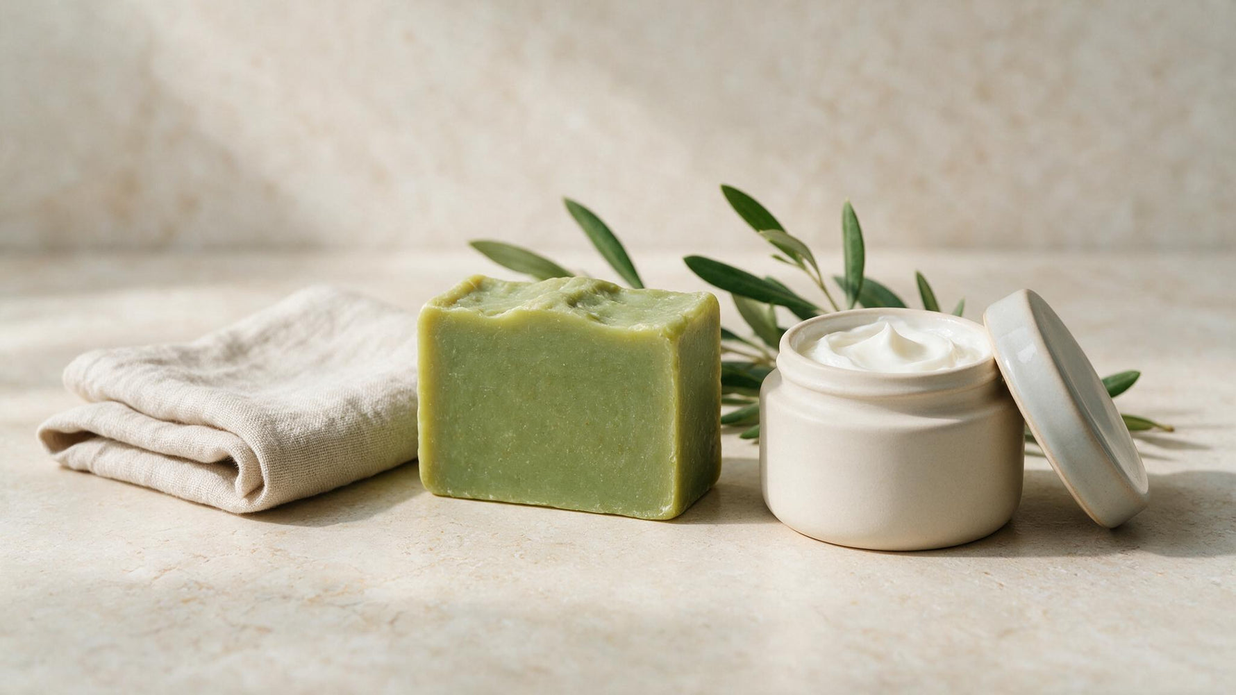 Natural additive-free skincare: an olive-oil soap bar, a simple cream jar and fresh olive branches on stone